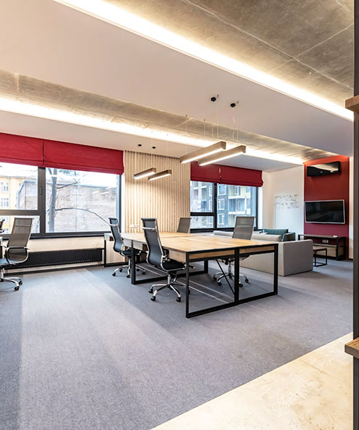 Collaborative office workspace interior in Dubai, featuring a large wooden desk with ergonomic chairs, a cozy sofa, red blinds, and modern lighting with a wall-mounted TV.