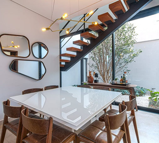 Modern residential dining area in Dubai, featuring a glossy white table with brown leather chairs, a contemporary staircase, decorative mirrors, and large windows with a view of a garden.