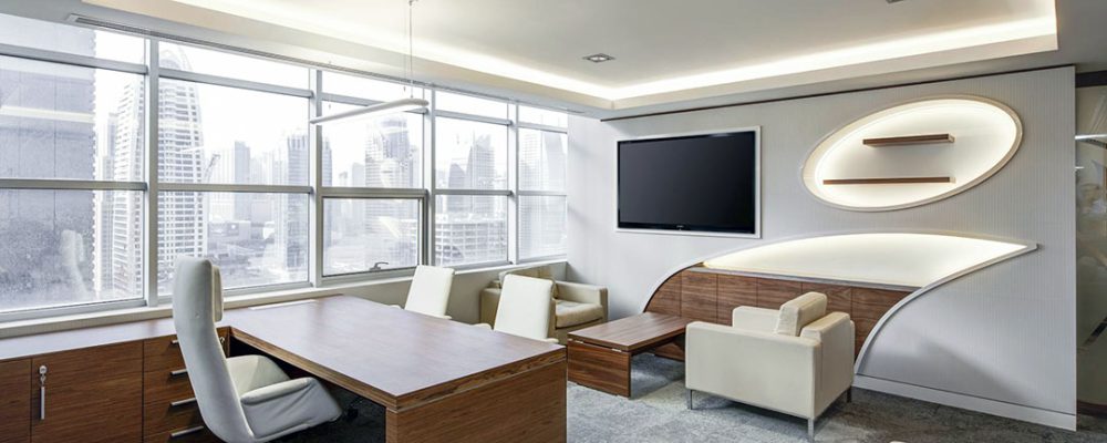 Executive office interior in Dubai, featuring a modern desk, white chairs, a cozy seating area, a wall-mounted TV, and large windows with panoramic city views.