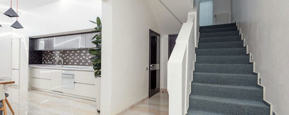 Modern residential interior in Dubai, featuring a sleek white kitchen with a tiled backsplash and a contemporary staircase with grey carpet, illuminated by pendant lighting and natural light.
